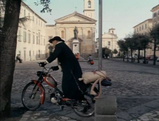 Il prete parte a bordo del suo ciclomotore, sullo sfondo la chiesa dell'Annunziata
